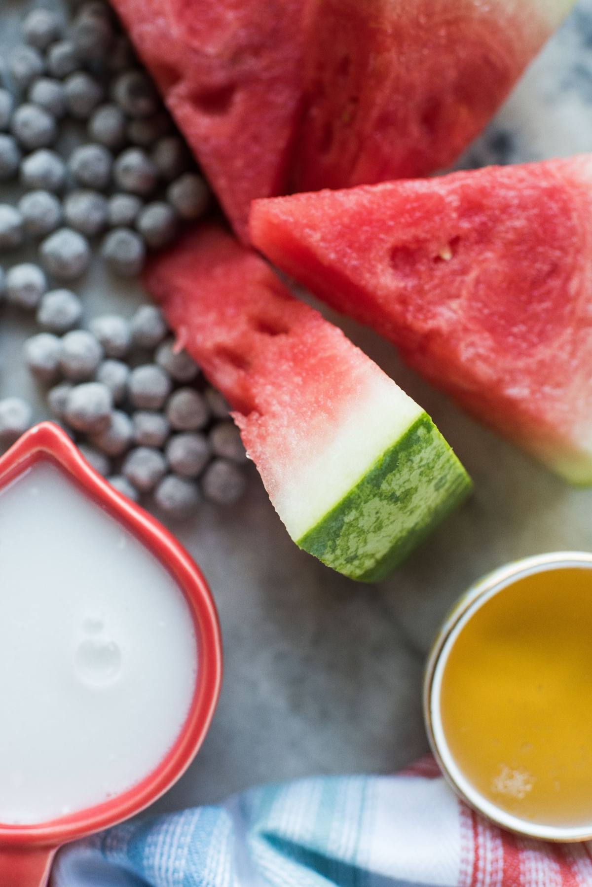 Watermelon slices, frozen blueberries, a pitcher of milk, and watermelon boba popsicles sit beside a bowl of orange liquid on the table.