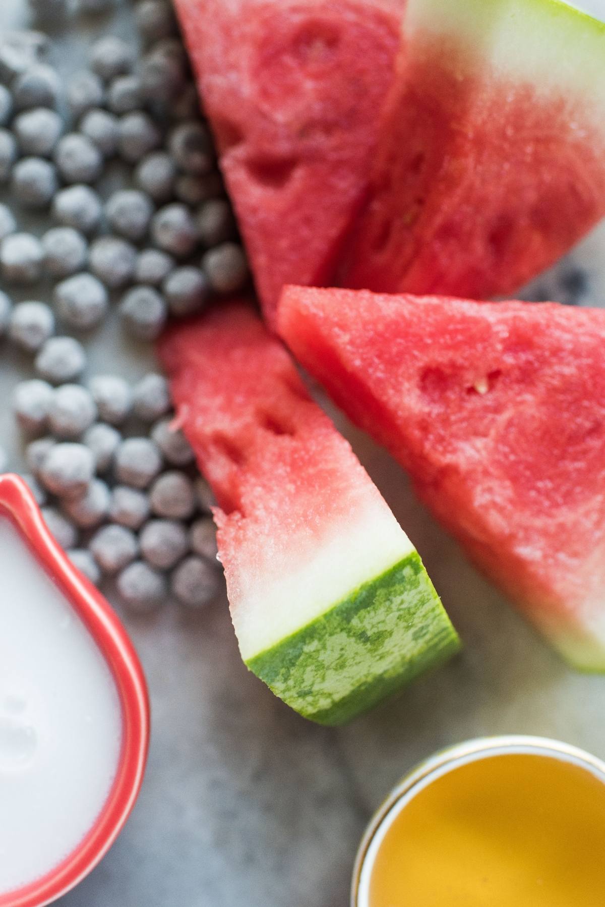 Slices of watermelon, frozen blueberries, coconut milk, and honey arranged on a light surface—perfect ingredients for refreshing watermelon boba popsicles.