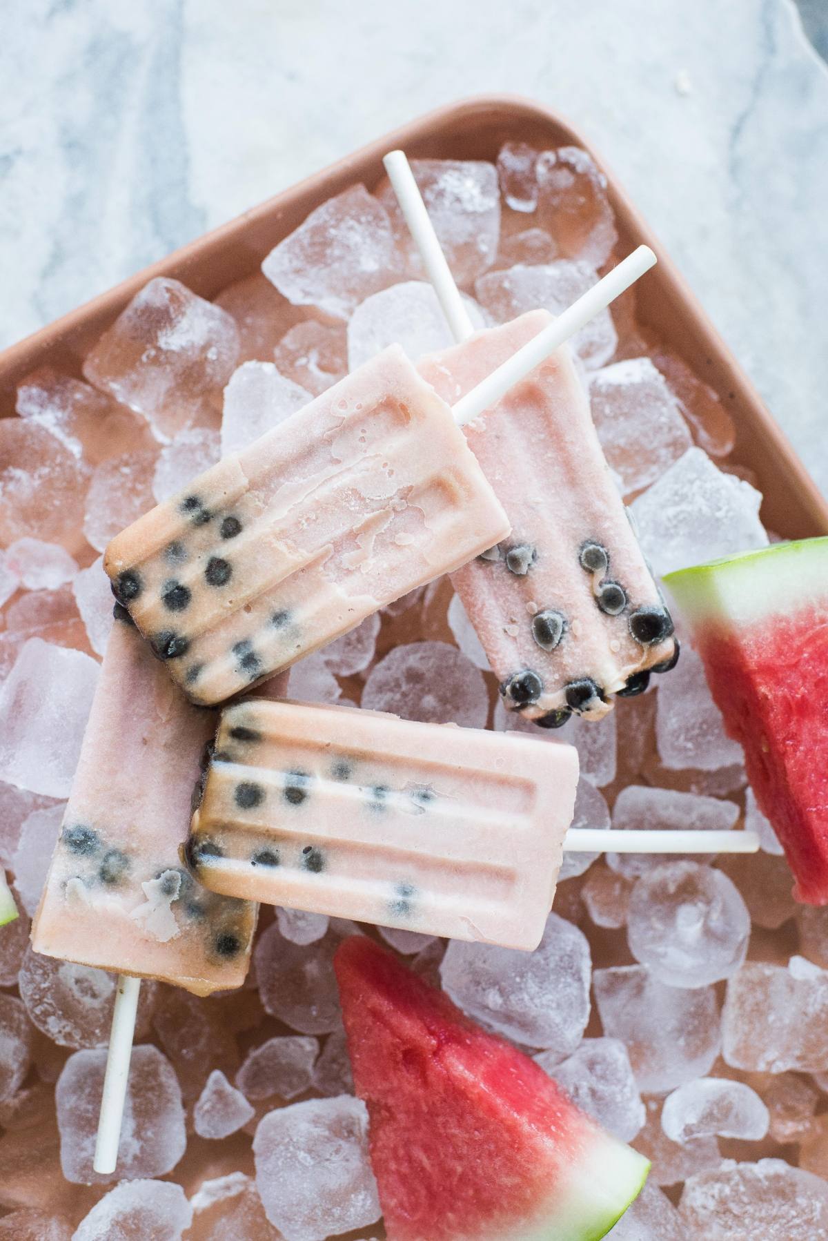 Three pink boba popsicles with black tapioca pearls rest on ice, next to juicy watermelon slices on a tray.