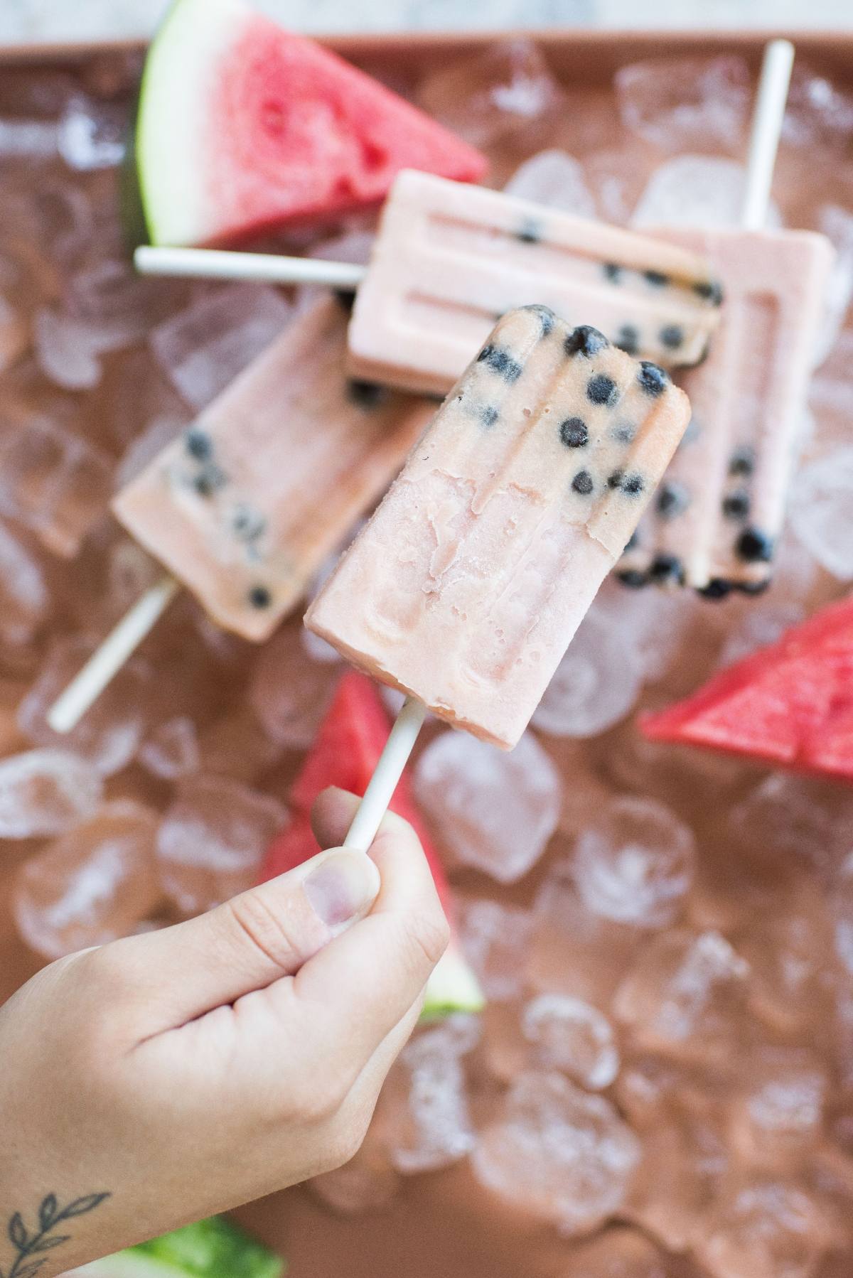 A hand holds a delicious watermelon boba popsicle with black seeds, over a tray of ice and fresh watermelon slices.