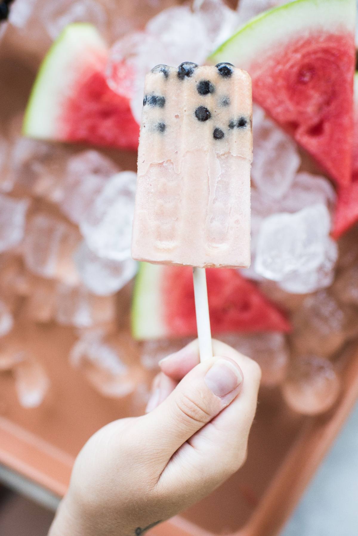 Hand holding a boba popsicle with black tapioca pearls, surrounded by ice and watermelon slices in the background.