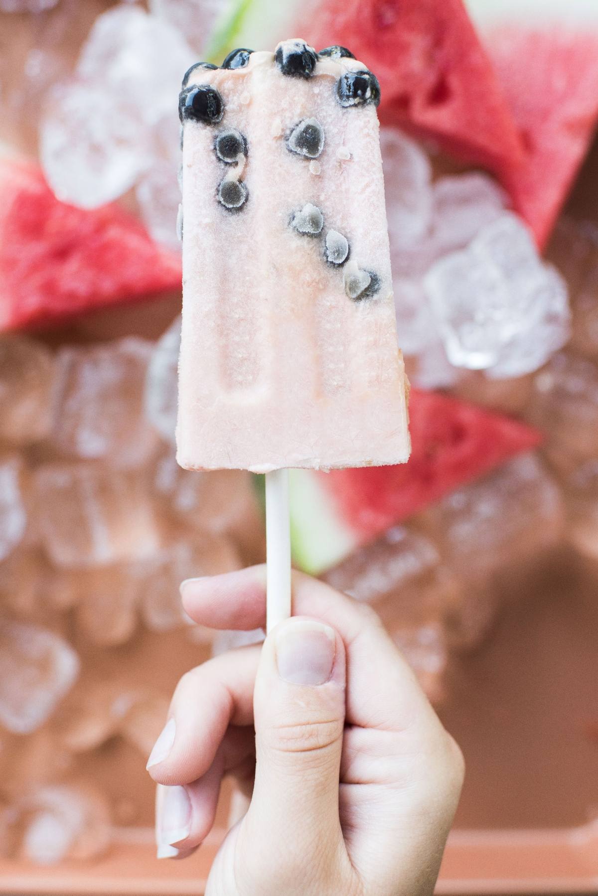 A hand holding a pink watermelon boba popsicle with black seeds, with watermelon slices and ice in the background.