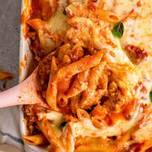 Baked mostaccioli pasta with melted cheese and basil leaves, served with a pink-handled spoon in a casserole dish.