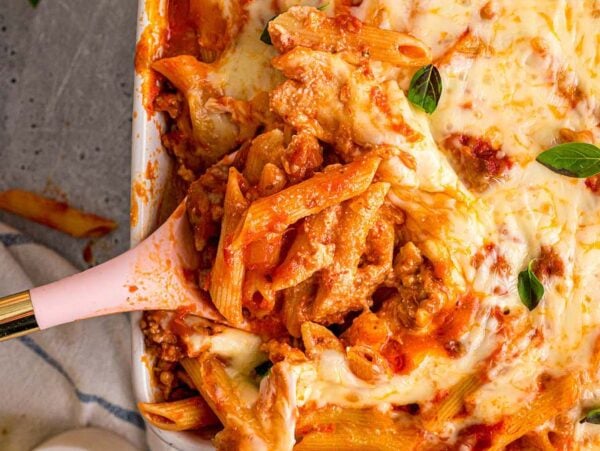 Baked mostaccioli pasta with melted cheese and basil leaves, served with a pink-handled spoon in a casserole dish.