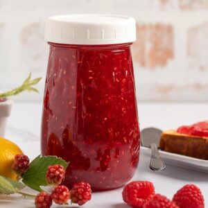 A jar of raspberry jam sits on a white table, surrounded by fresh raspberries, lemons, and slices of toast.