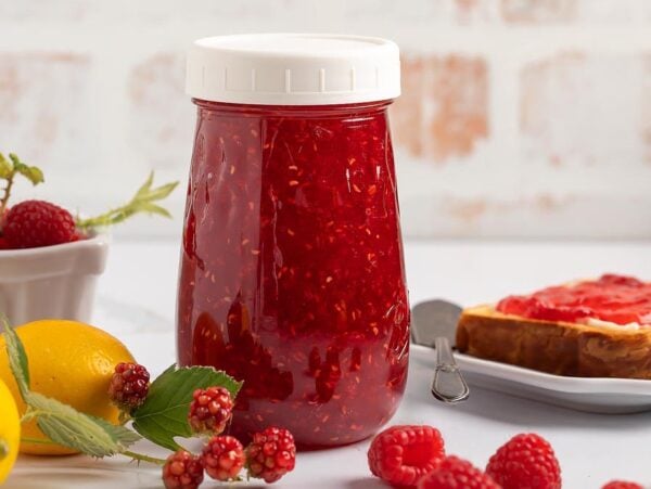 A jar of raspberry jam sits on a white table, surrounded by fresh raspberries, lemons, and slices of toast.
