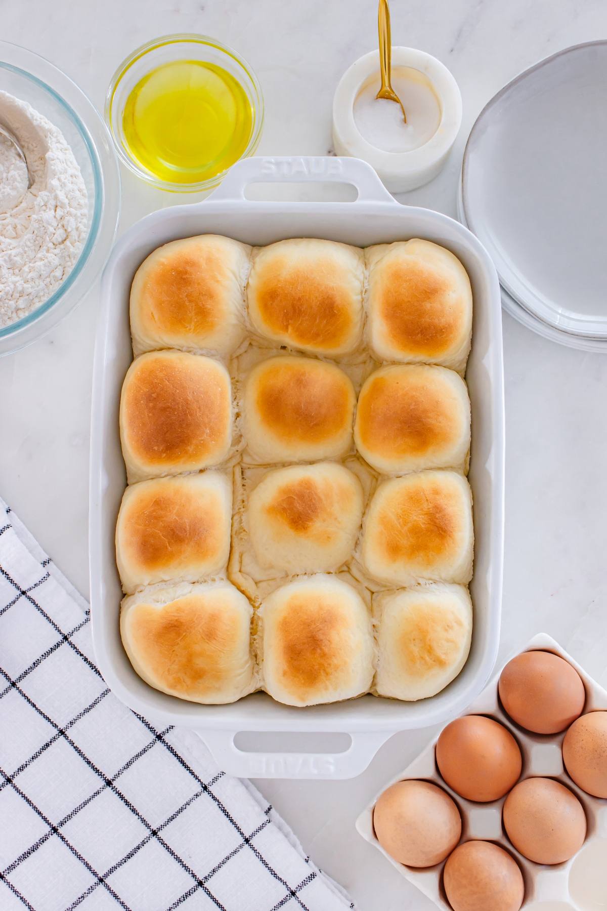A baking dish with twelve golden dinner rolls, surrounded by eggs, flour, oil, and plates on a countertop.