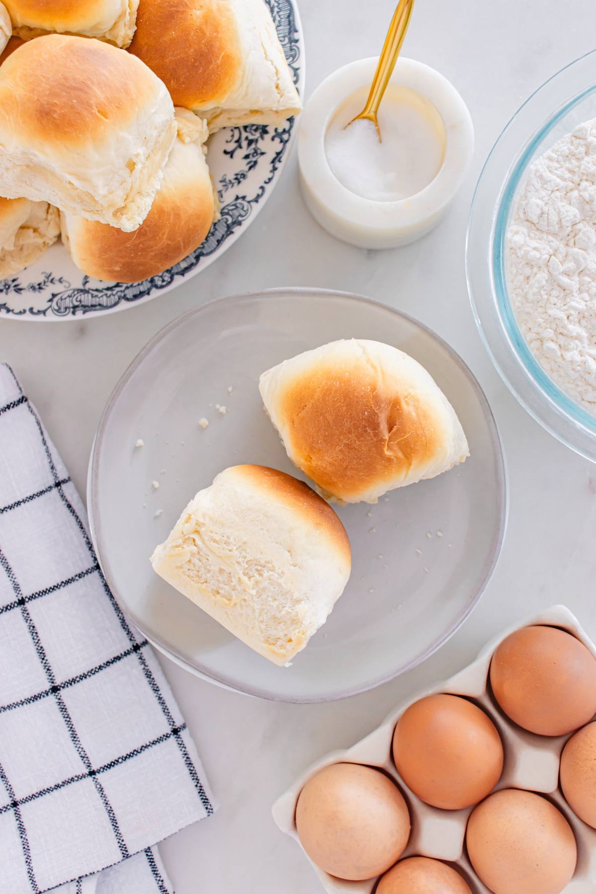 Two dinner rolls on a plate, surrounded by eggs, flour, sugar, and more rolls on a patterned plate.