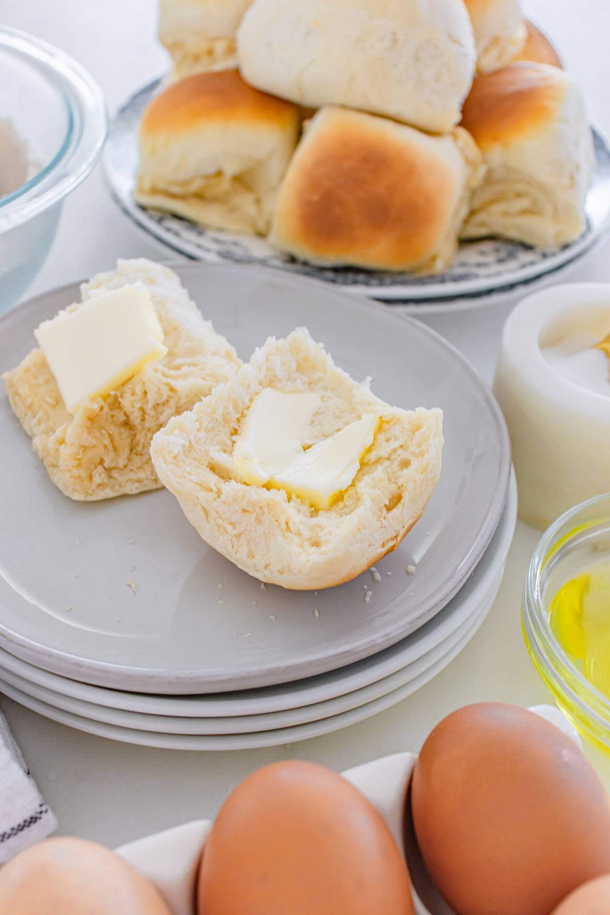 A biscuit split open with butter, surrounded by eggs, rolls, and a bowl of oil on a table.