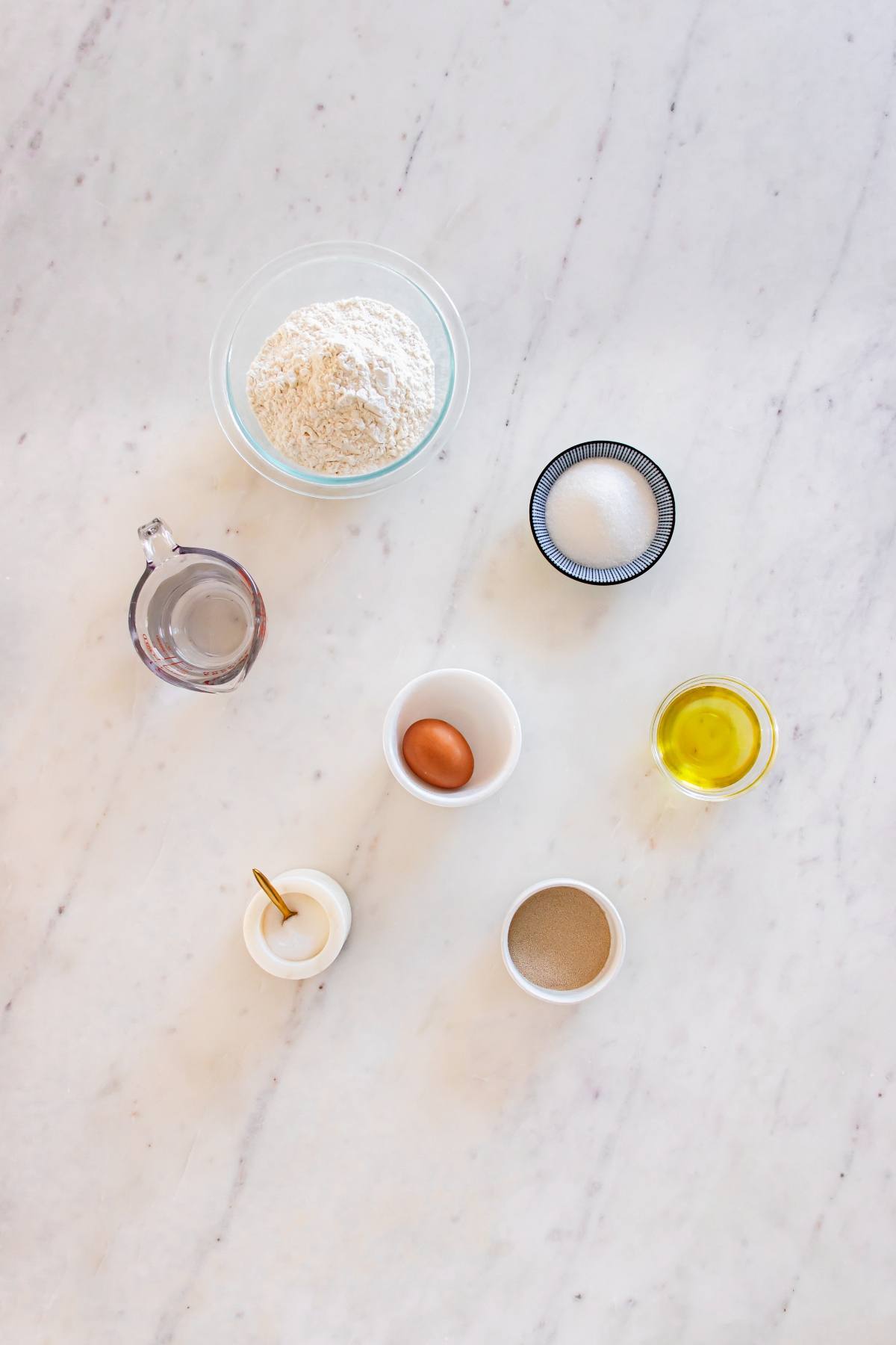 Bowl of flour, sugar, egg, oil, water, salt, and yeast on a white marble surface.