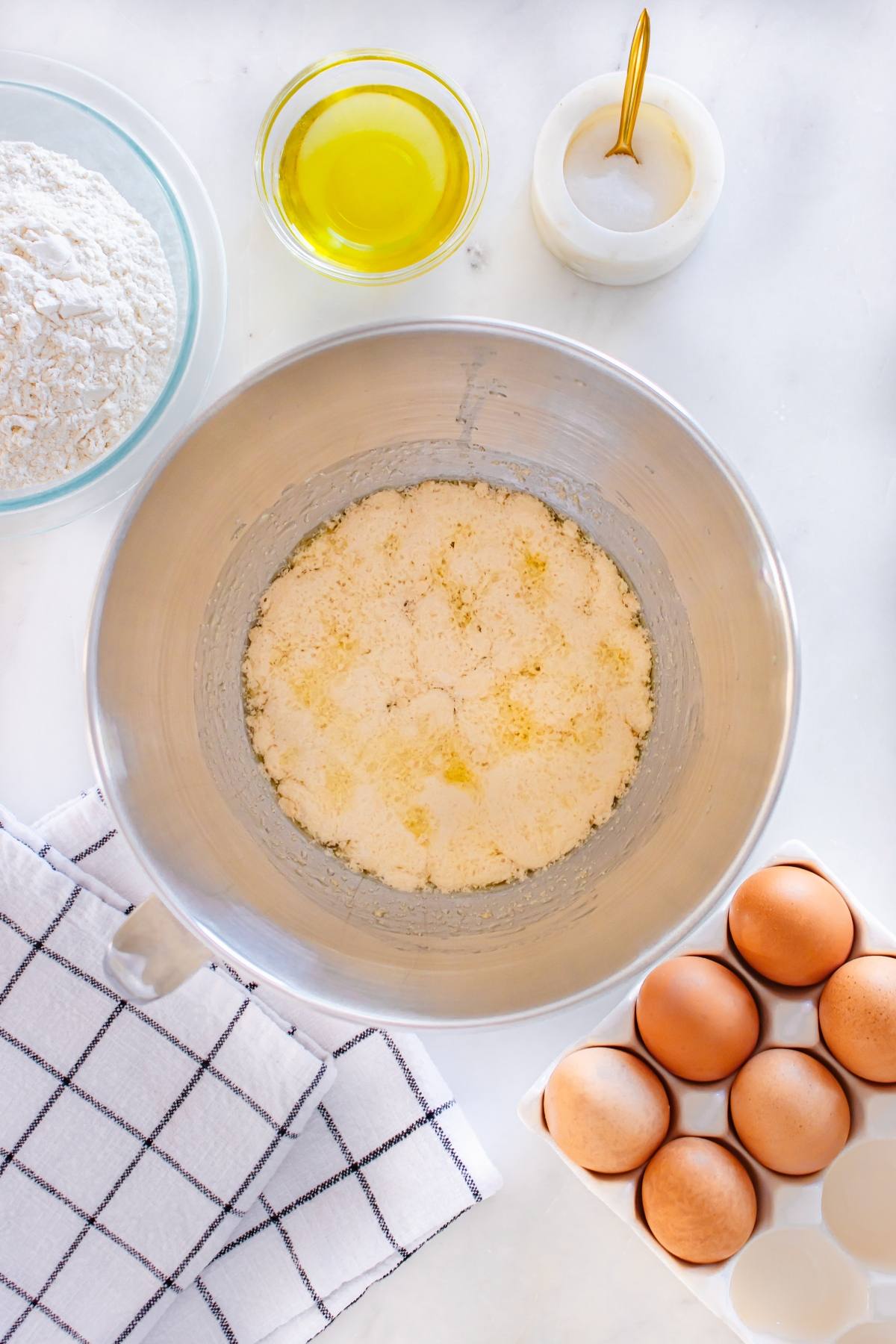 A mixing bowl with wet ingredients, eggs in a carton, flour, oil, and salt arranged on a countertop with a towel.