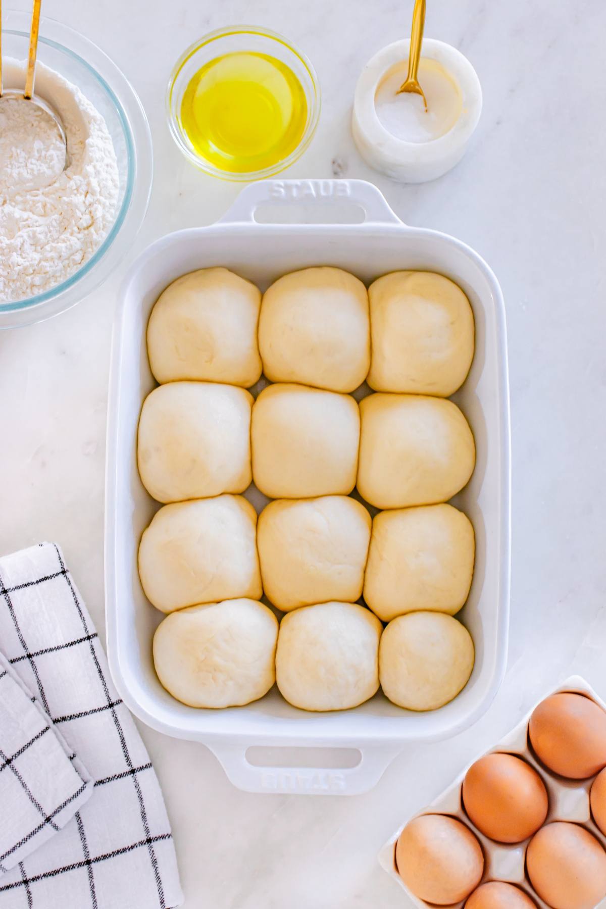 Twelve dough balls in a white baking dish, surrounded by baking ingredients on a white counter.