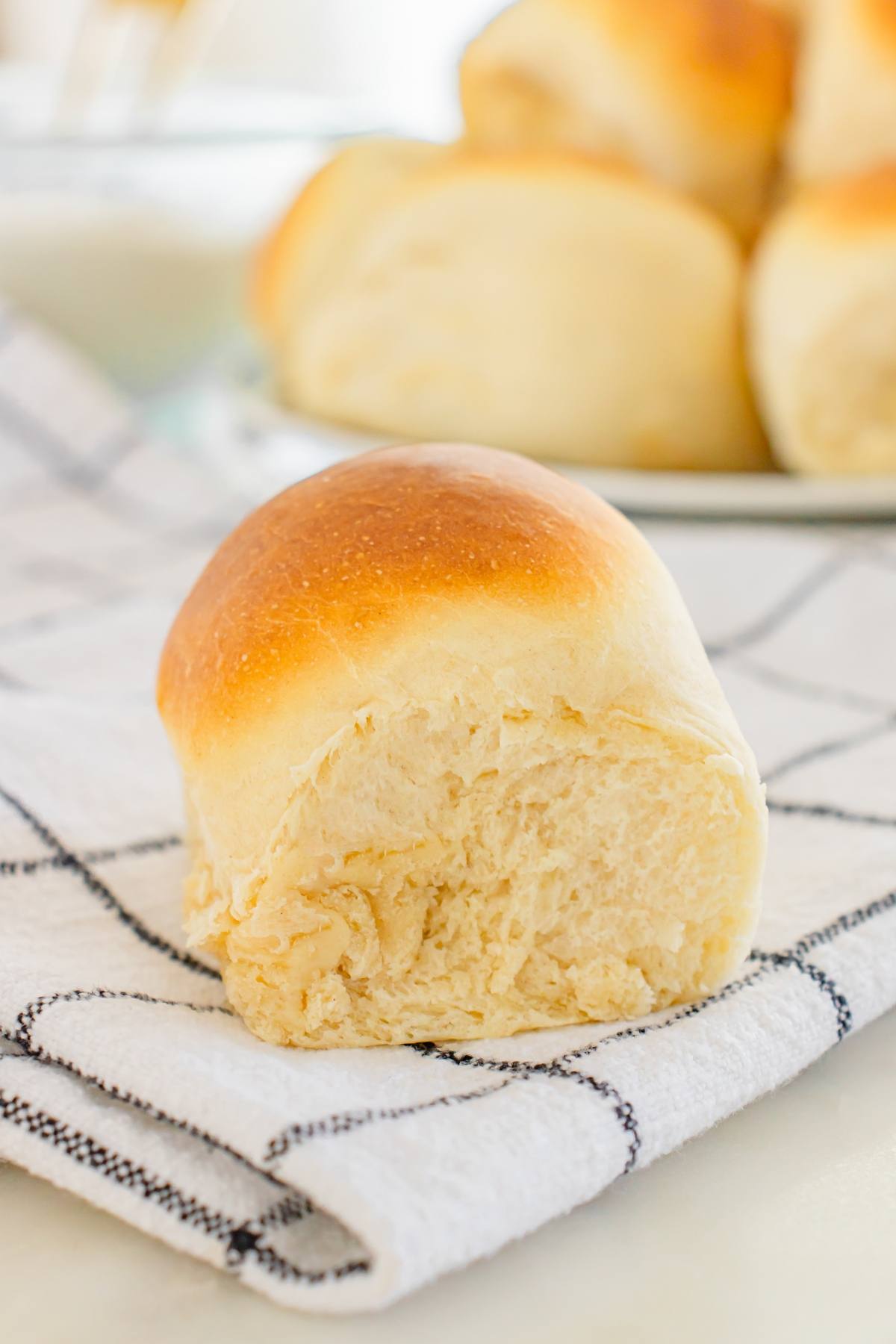 A soft, golden-brown dinner roll with a bite taken out, resting on a white checkered cloth.