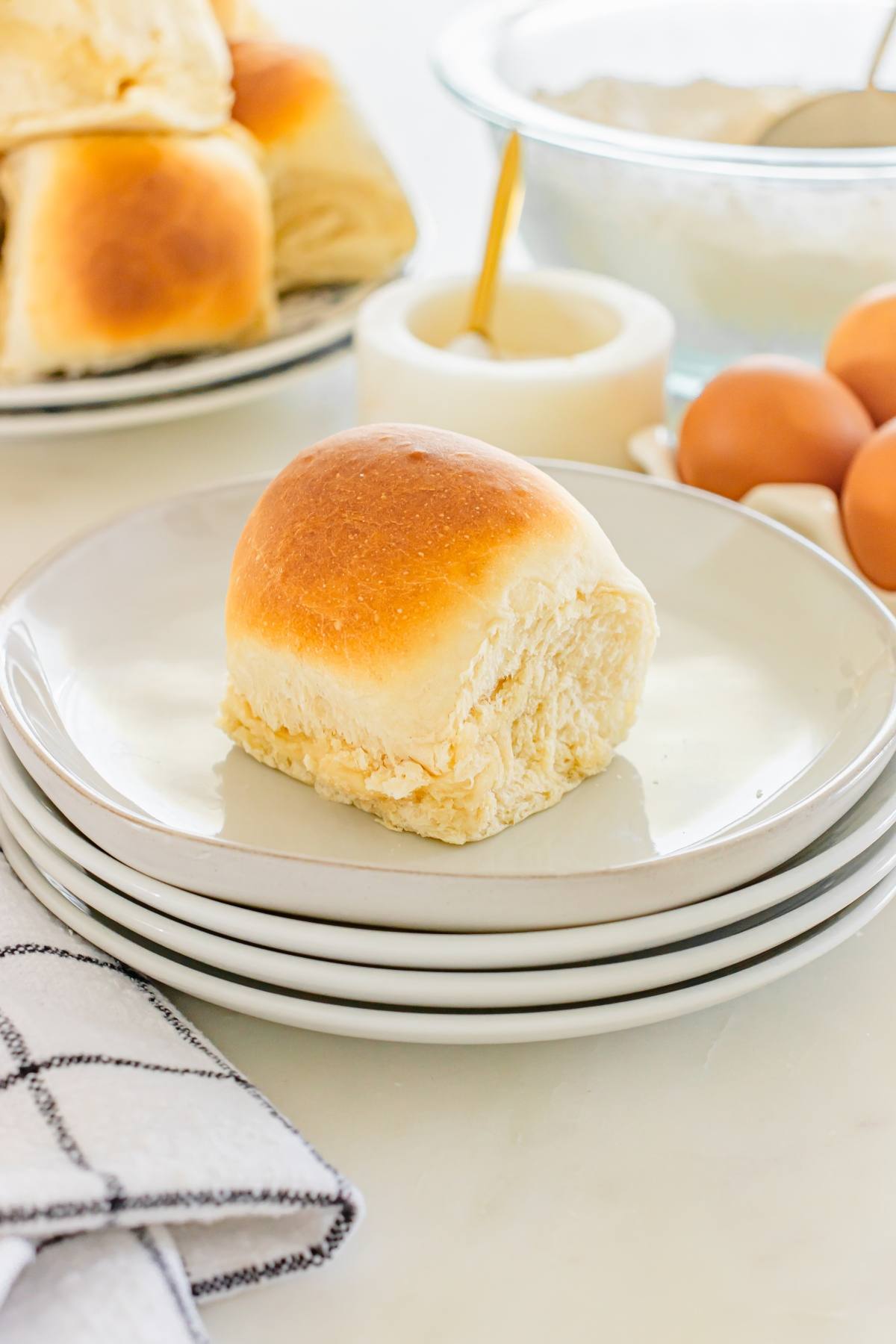 A golden-brown dinner roll sits on a stack of white plates, with eggs and more rolls in the background.