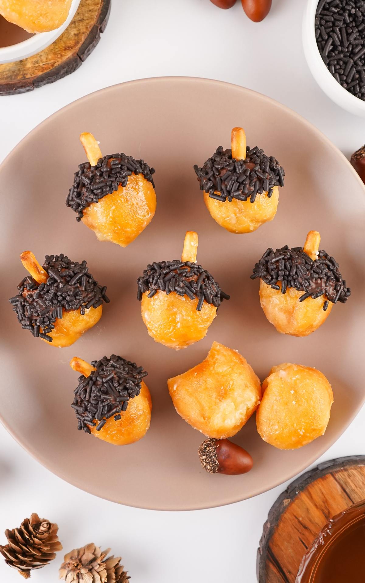 Seven donut holes decorated like acorns with chocolate sprinkles and pretzel sticks on a beige plate.