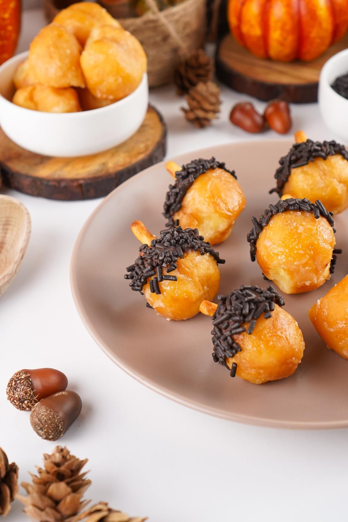 Golden donut holes with chocolate sprinkles on a plate, styled like acorns with autumn decorations nearby.