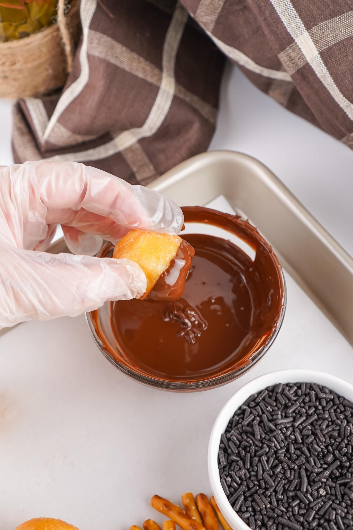 Gloved hand dipping a pastry into melted chocolate, with chocolate sprinkles in a bowl nearby.
