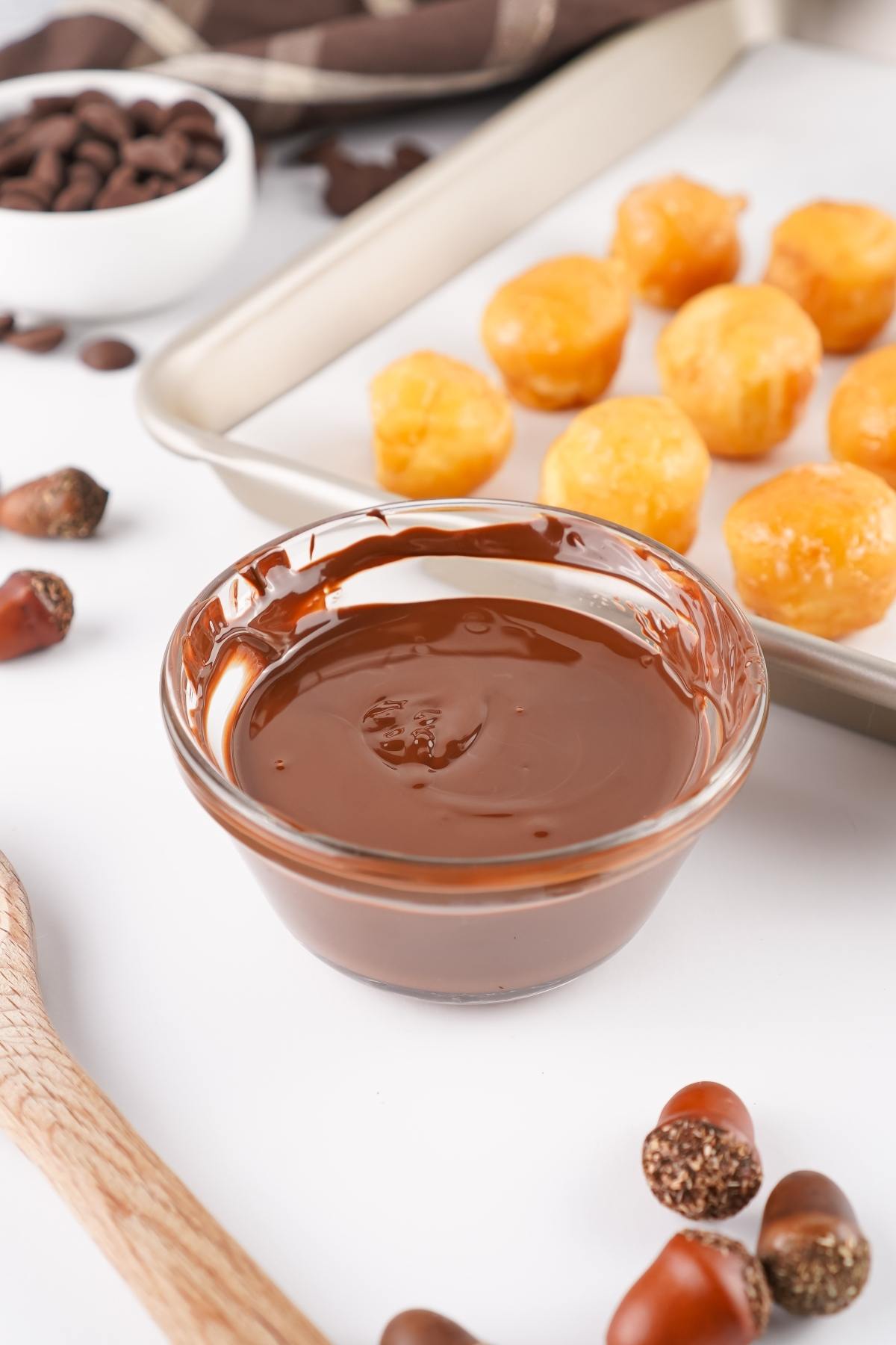 A glass bowl of melted chocolate with dough balls on a tray and scattered acorns nearby.