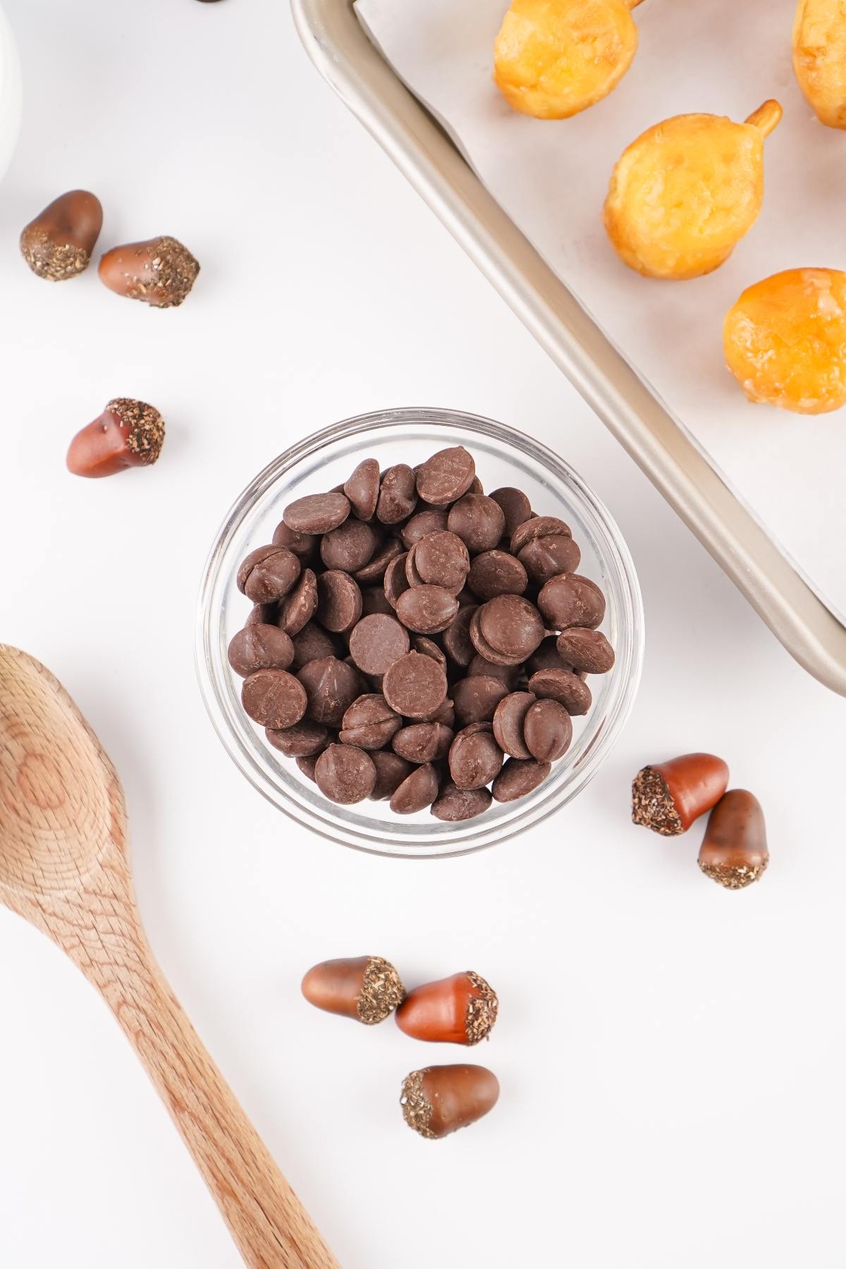A bowl of chocolate chips, mini acorn candies, doughnut holes, and a wooden spoon on a white surface.