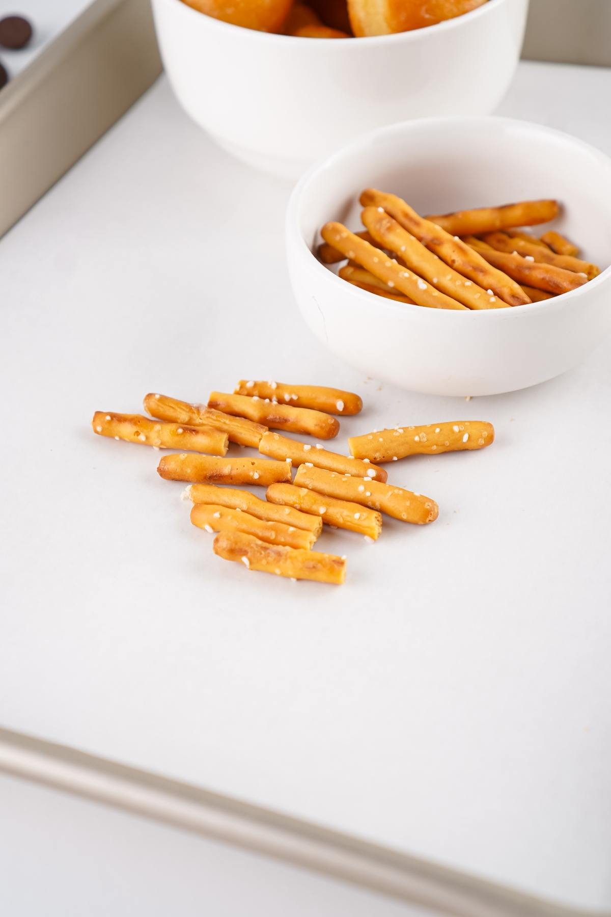 Pretzel sticks scattered on a white surface, with more pretzel sticks in a white bowl nearby.