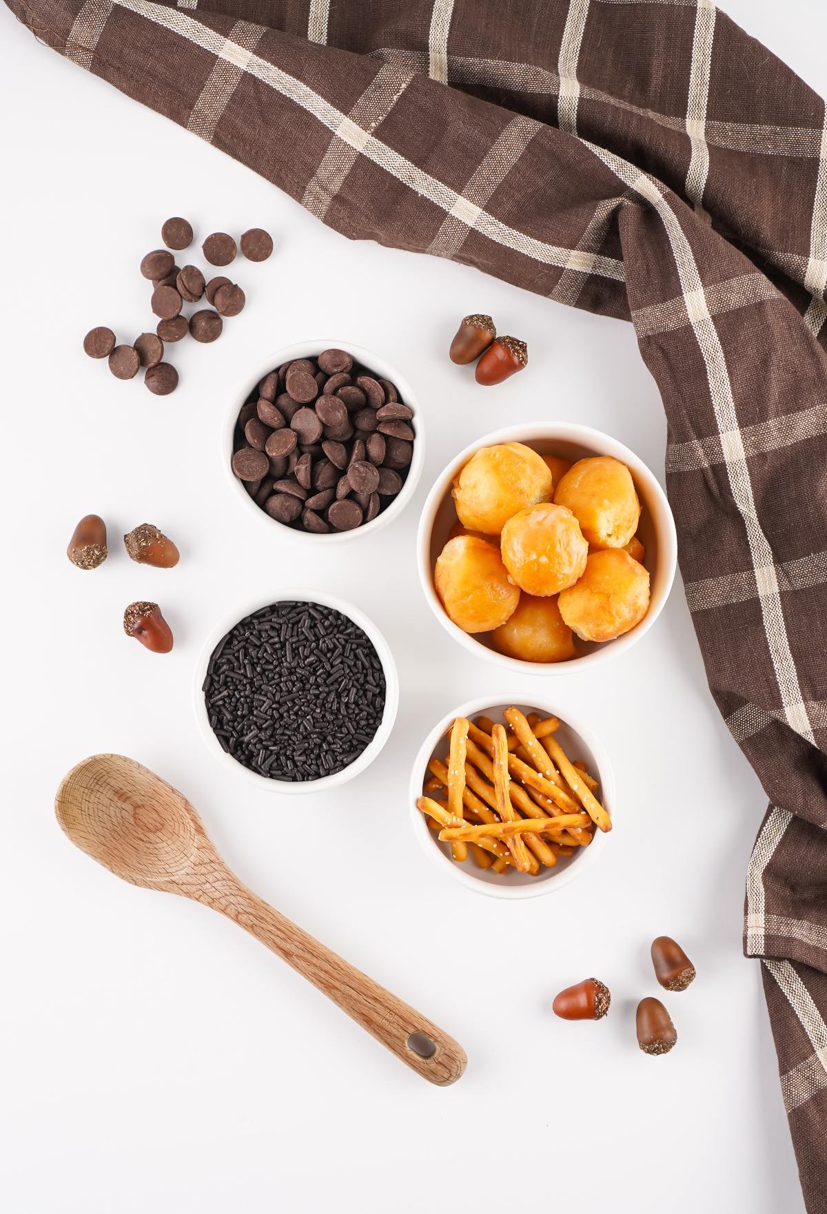 Wooden spoon, chocolate chips, pretzel sticks, black sprinkles, pastry balls, acorns, and a brown plaid cloth.