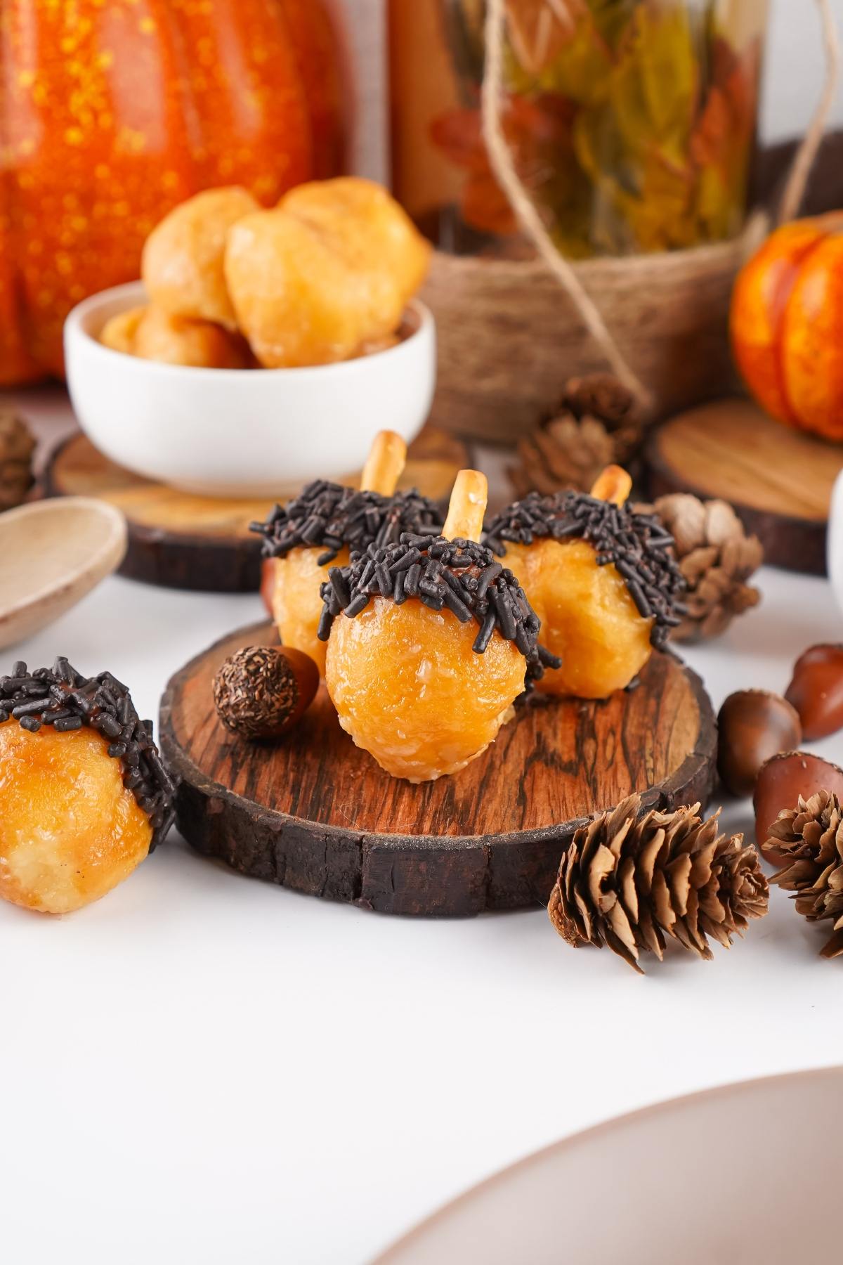 Three donut holes decorated as acorns with chocolate sprinkles and pretzel sticks, surrounded by autumn decor.