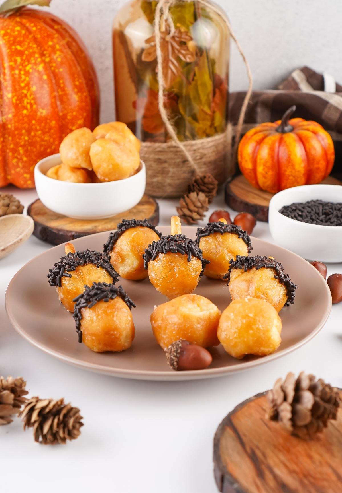Acorn-shaped donut holes with chocolate sprinkles on a plate, surrounded by autumn decorations and pumpkins.