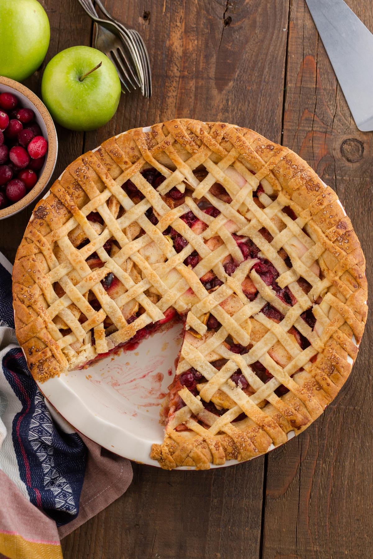 Lattice-topped pie with a slice missing, surrounded by green apples and cranberries on a wooden table.