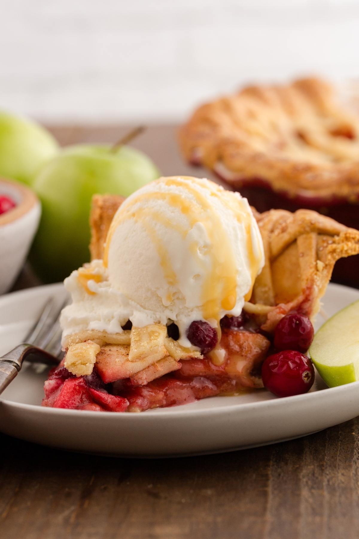A slice of apple pie with ice cream and caramel sauce on a plate, garnished with cranberries and apple slices.