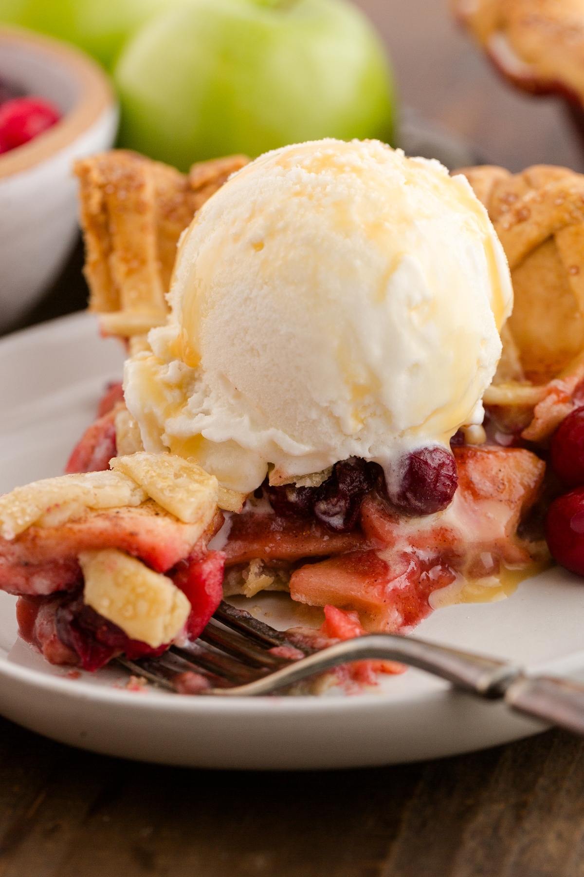 Slice of fruit pie with berries and apples, topped with vanilla ice cream, on a white plate with a fork.