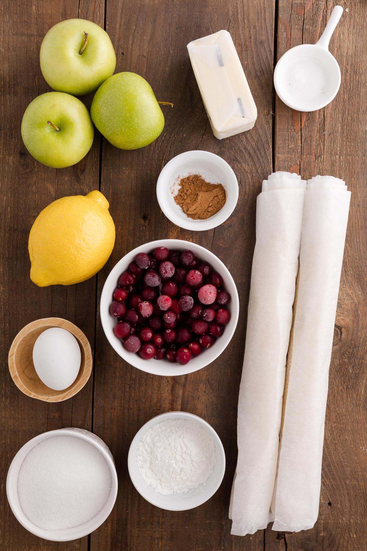 Ingredients for baking: apples, butter, sugar, lemon, egg, berries, cinnamon, flour, and pastry sheets on wood table.