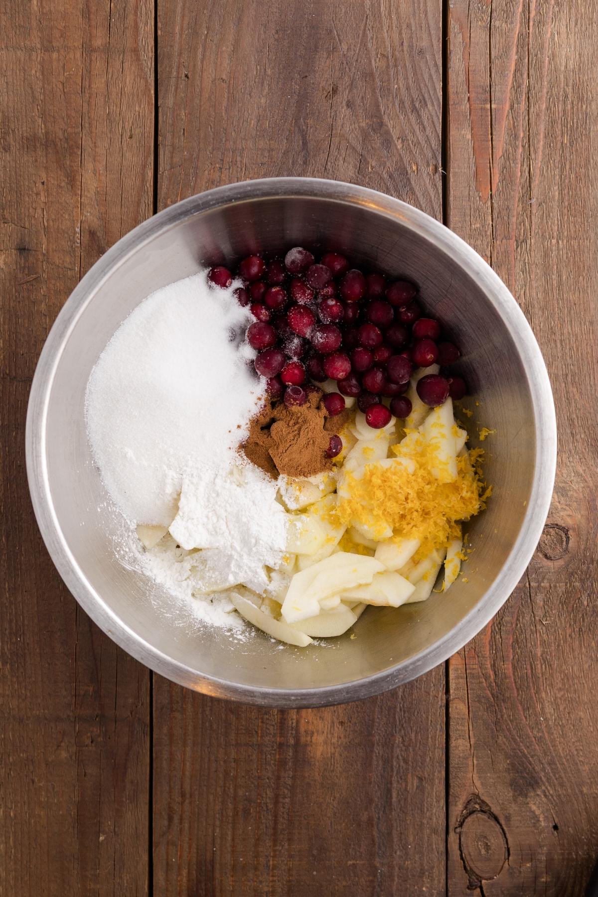 A metal bowl with cranberries, sliced apples, sugar, cinnamon, and lemon zest on a wooden surface.
