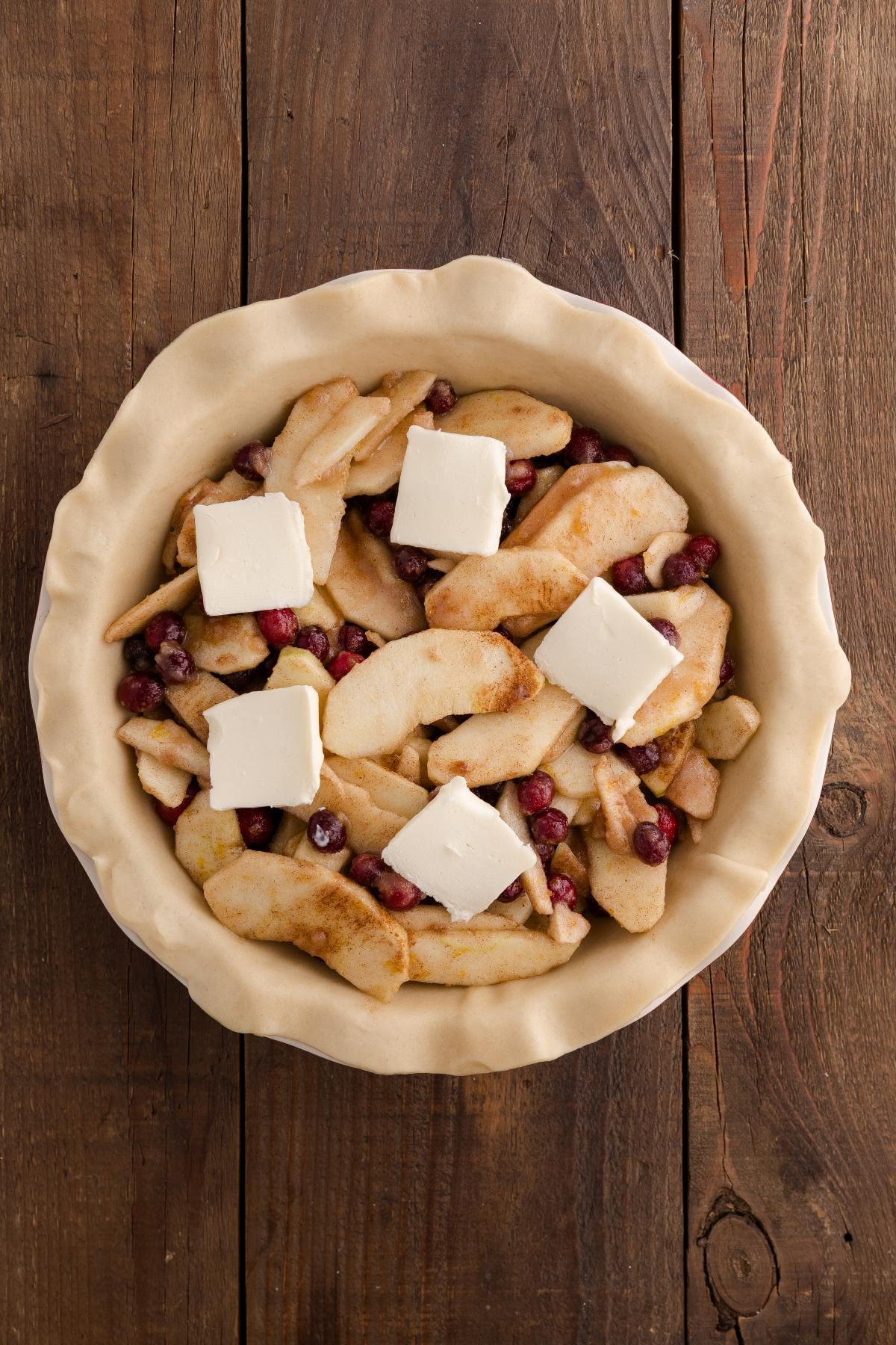 Unbaked pie with sliced apples, cranberries, and butter in a crust on a wooden table.