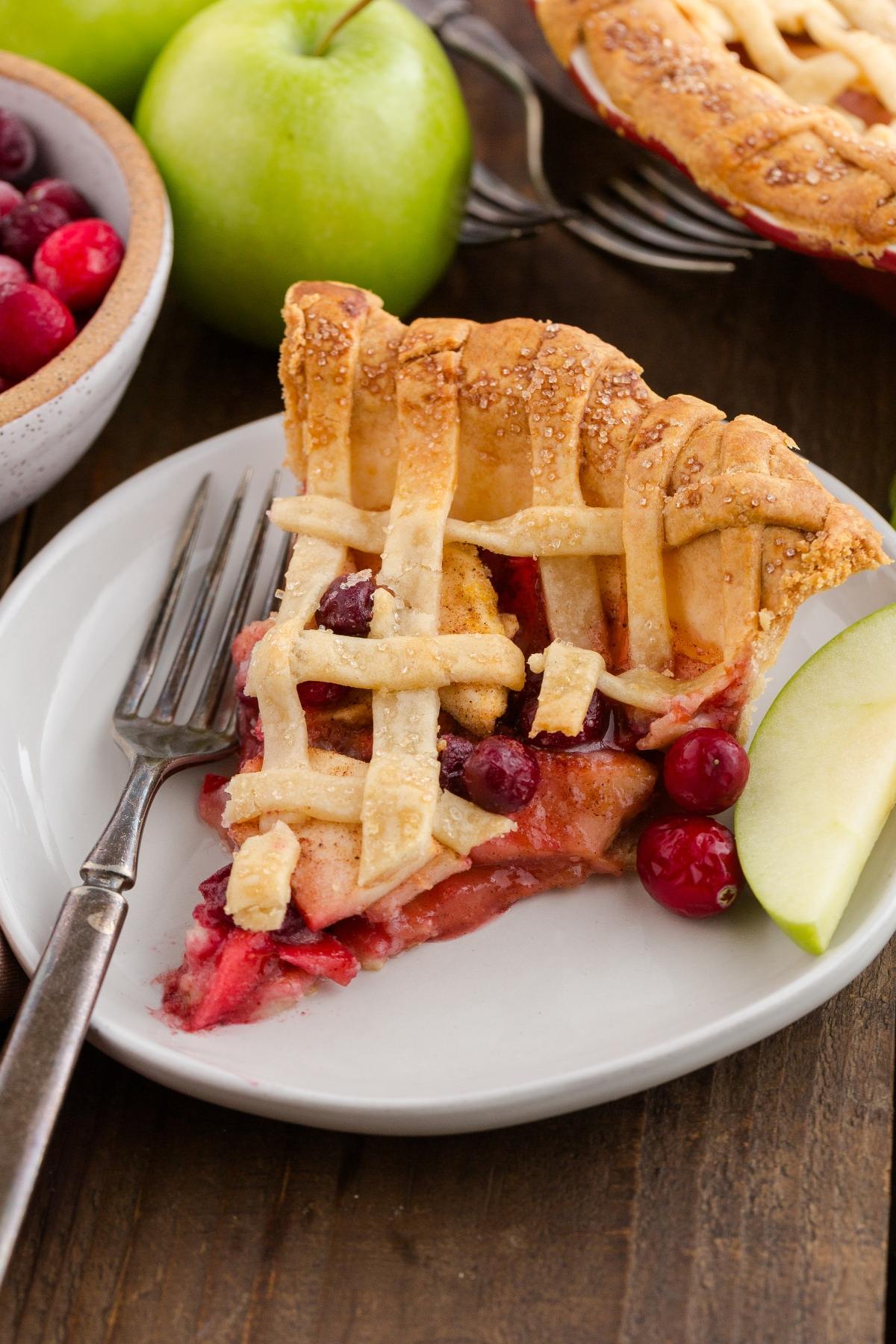 A slice of lattice-topped fruit pie with cranberries and apples on a plate, next to a fork and apple slice.
