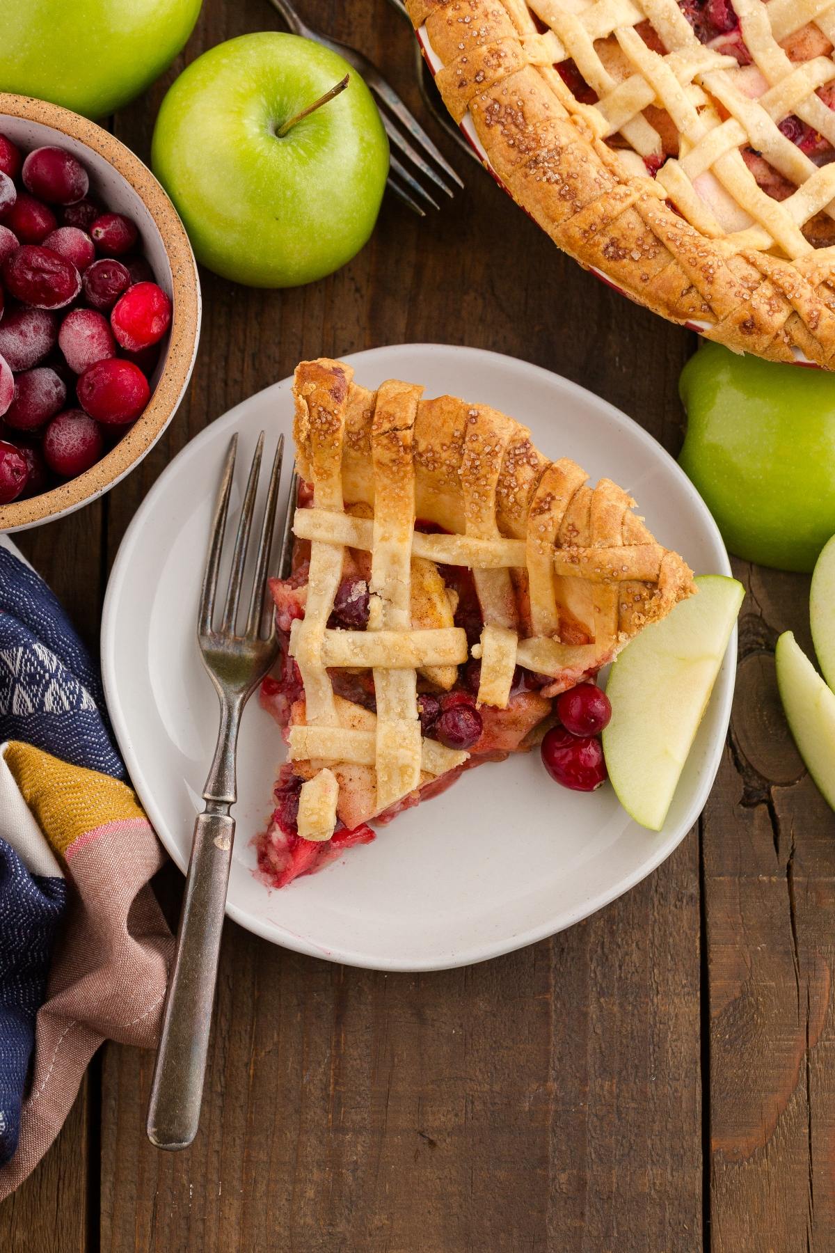 Slice of lattice-topped pie with cranberries on a plate, next to fresh apples and a bowl of cranberries.