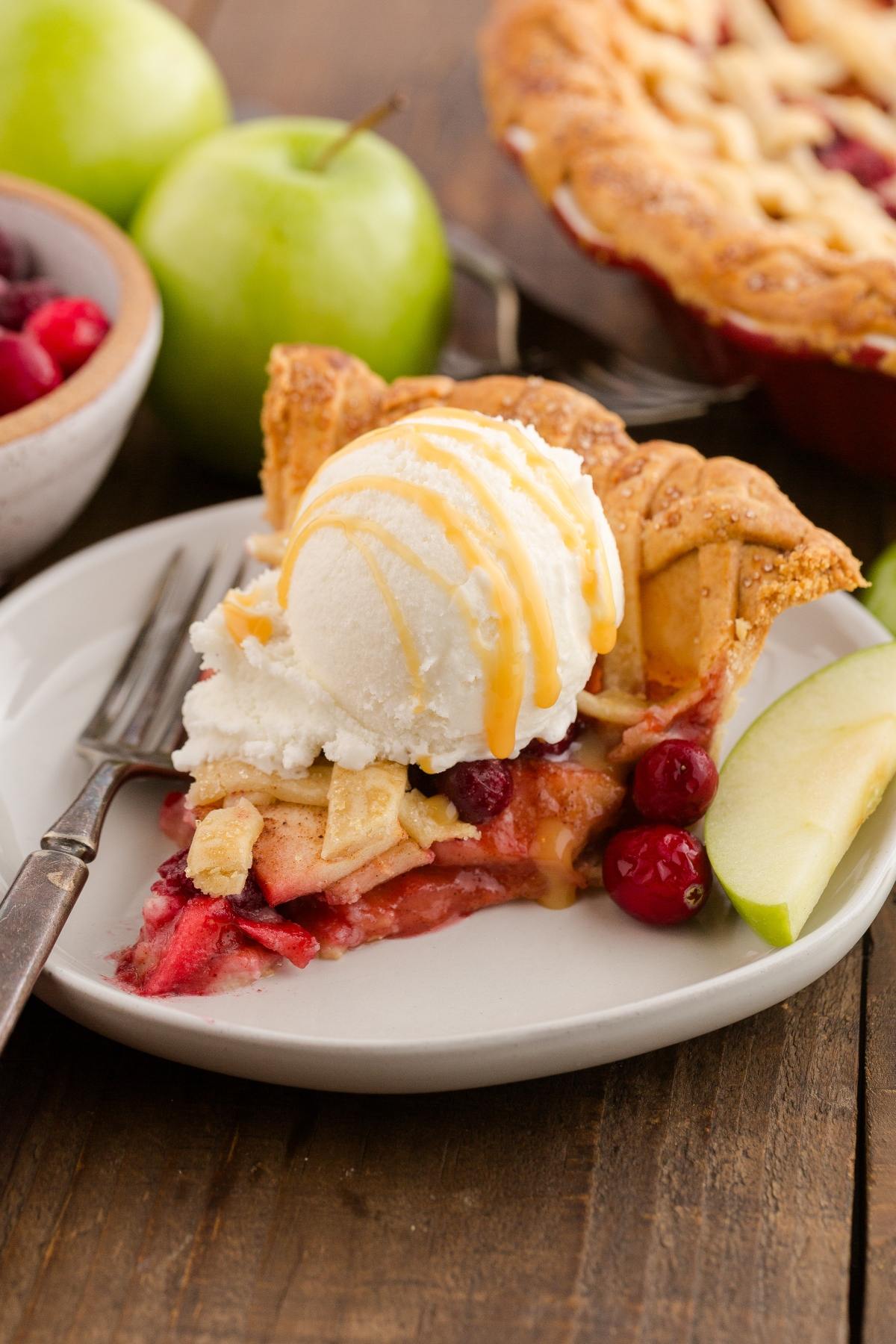A slice of fruit pie with ice cream and caramel drizzle on a plate, next to apple slices and a fork.