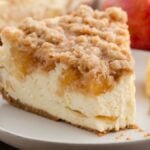 A slice of apple pie cheesecake on a white plate with fresh apples in the background.