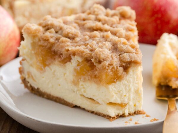 A slice of apple pie cheesecake on a white plate with fresh apples in the background.