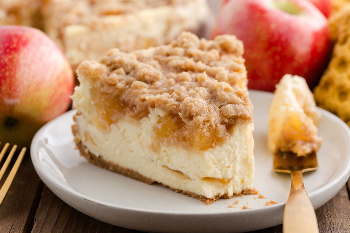 A slice of apple pie cheesecake on a white plate with fresh apples in the background.