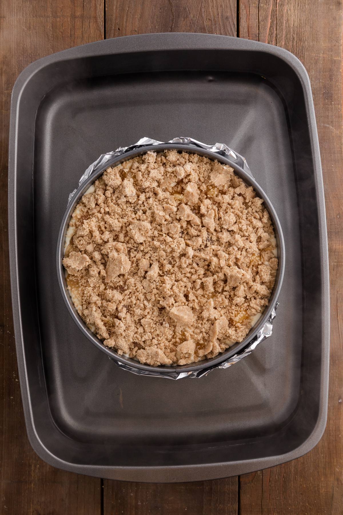 Round baking pan with crumbly topping inside a larger rectangular roasting pan on a wooden surface.