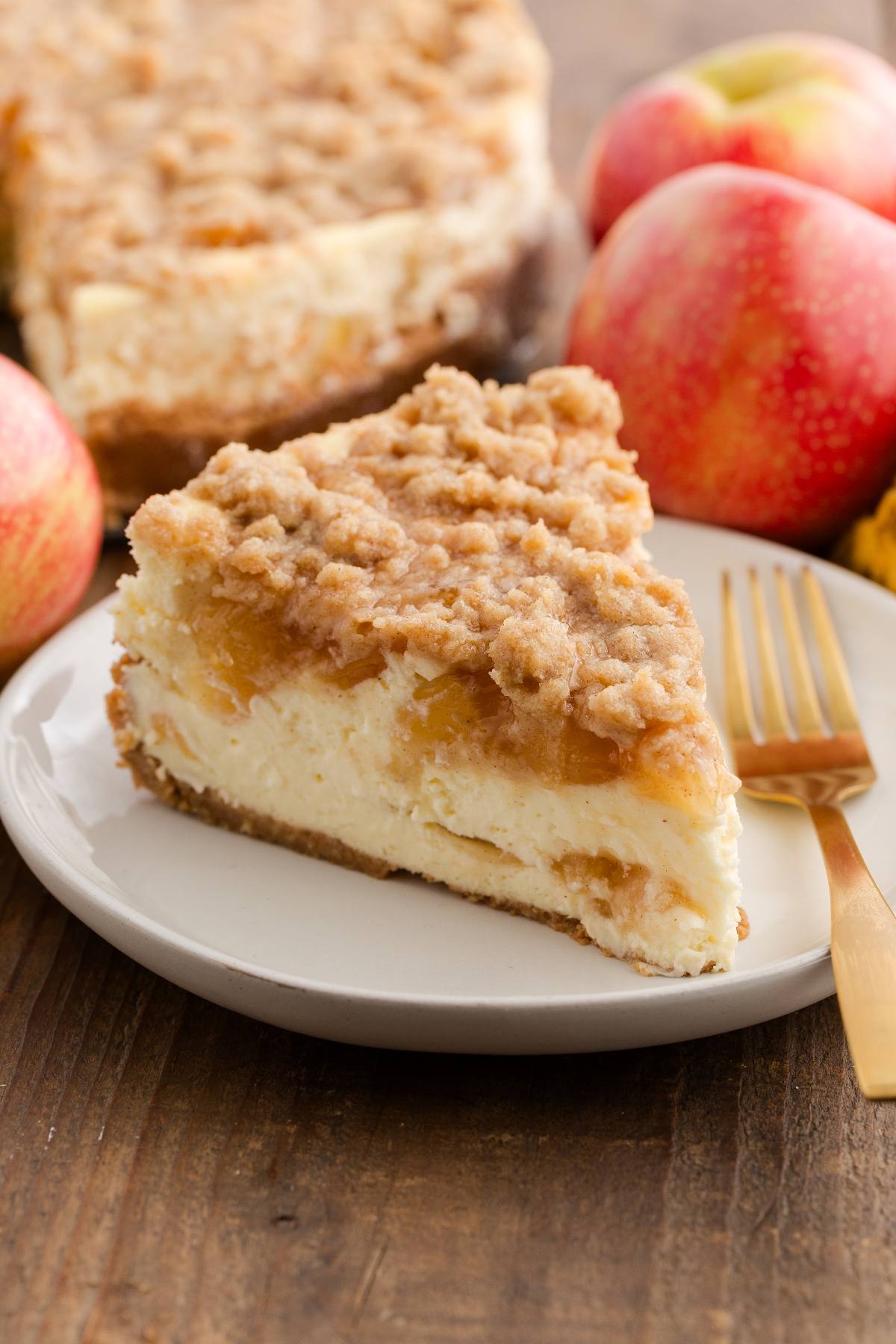 A slice of apple crumble cheesecake on a plate with apples and a fork beside it.