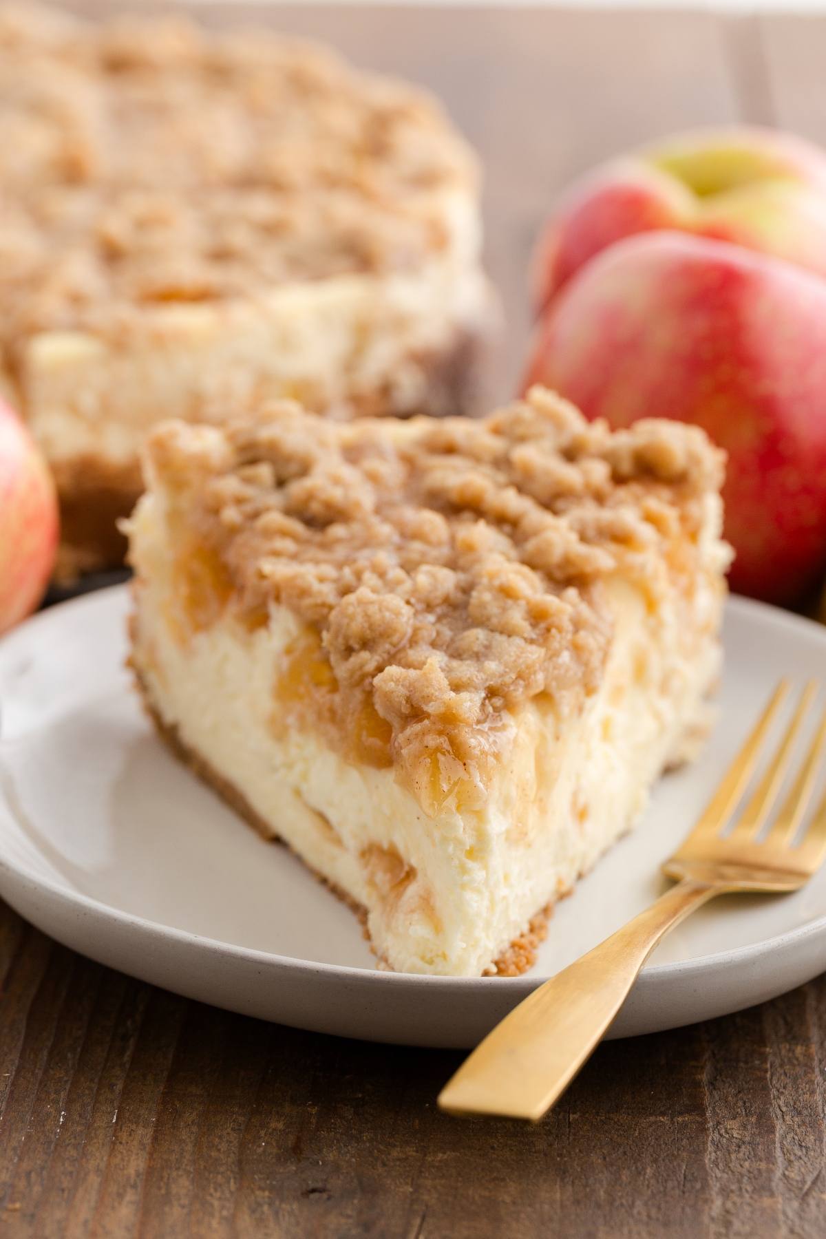 A slice of apple crumb cheesecake on a plate with apples and a gold fork in the background.