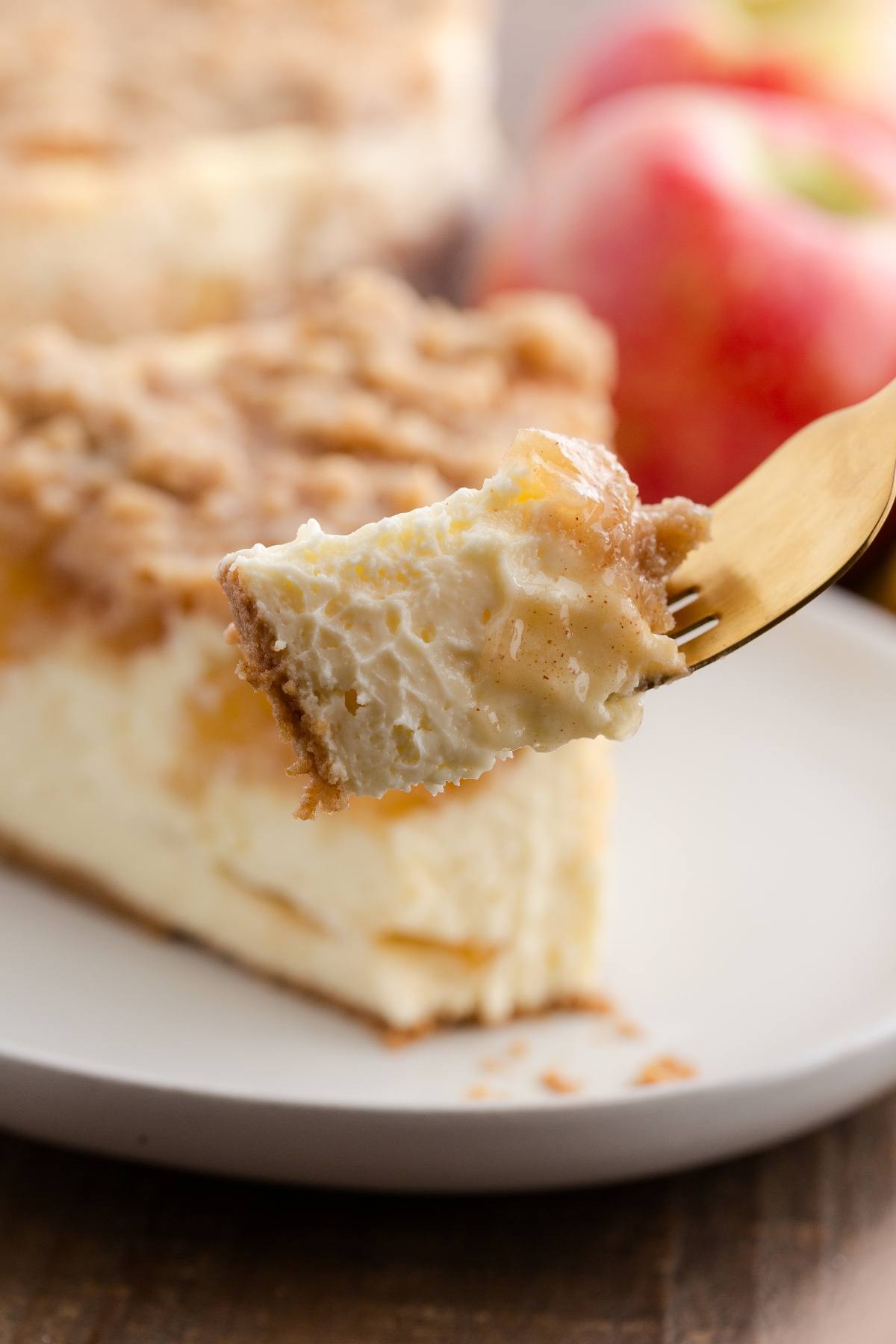 A fork holds a creamy bite of cheesecake with crumb topping; apples blurred in the background.