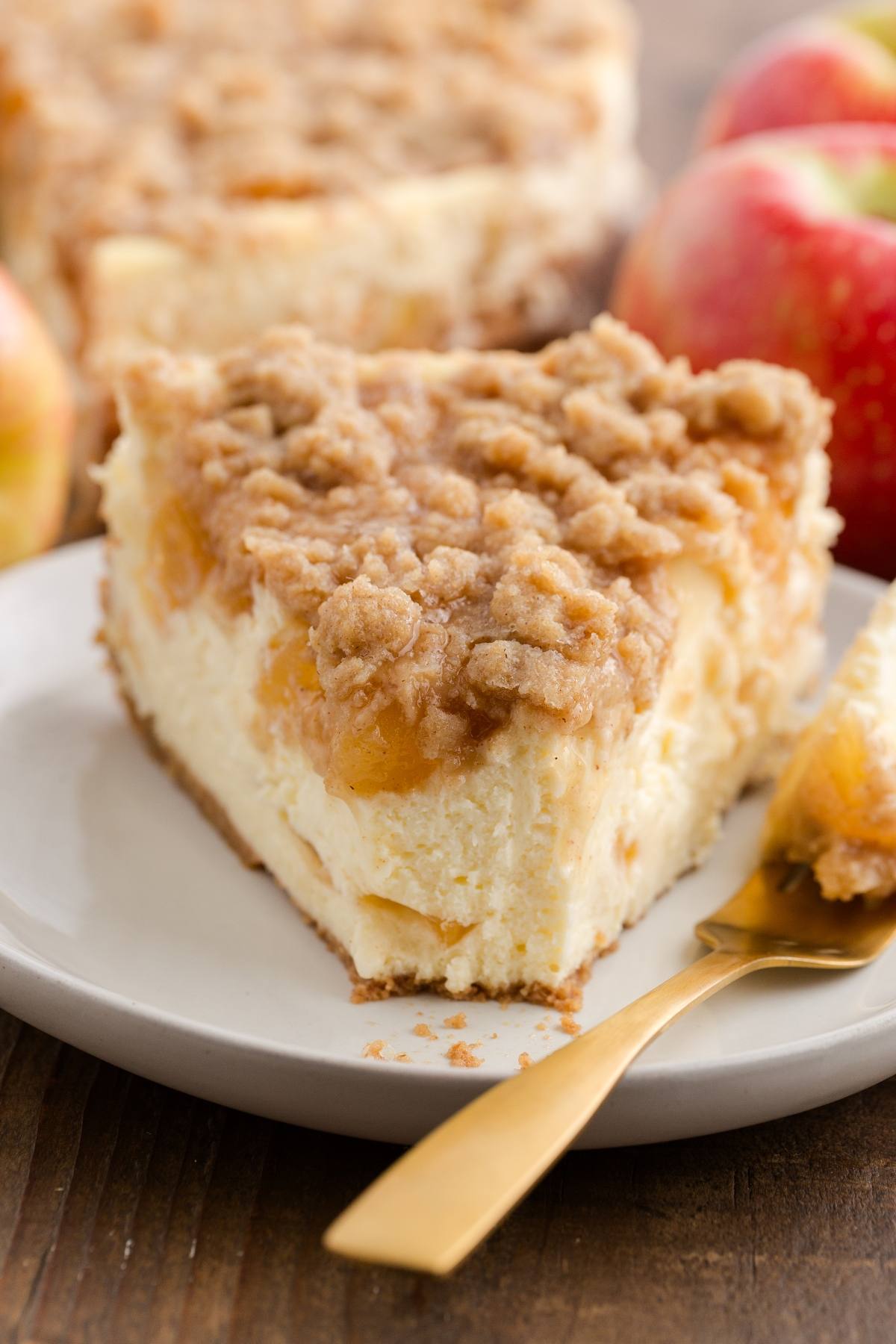 A slice of apple crumble cheesecake on a white plate with a gold fork, apples in the background.