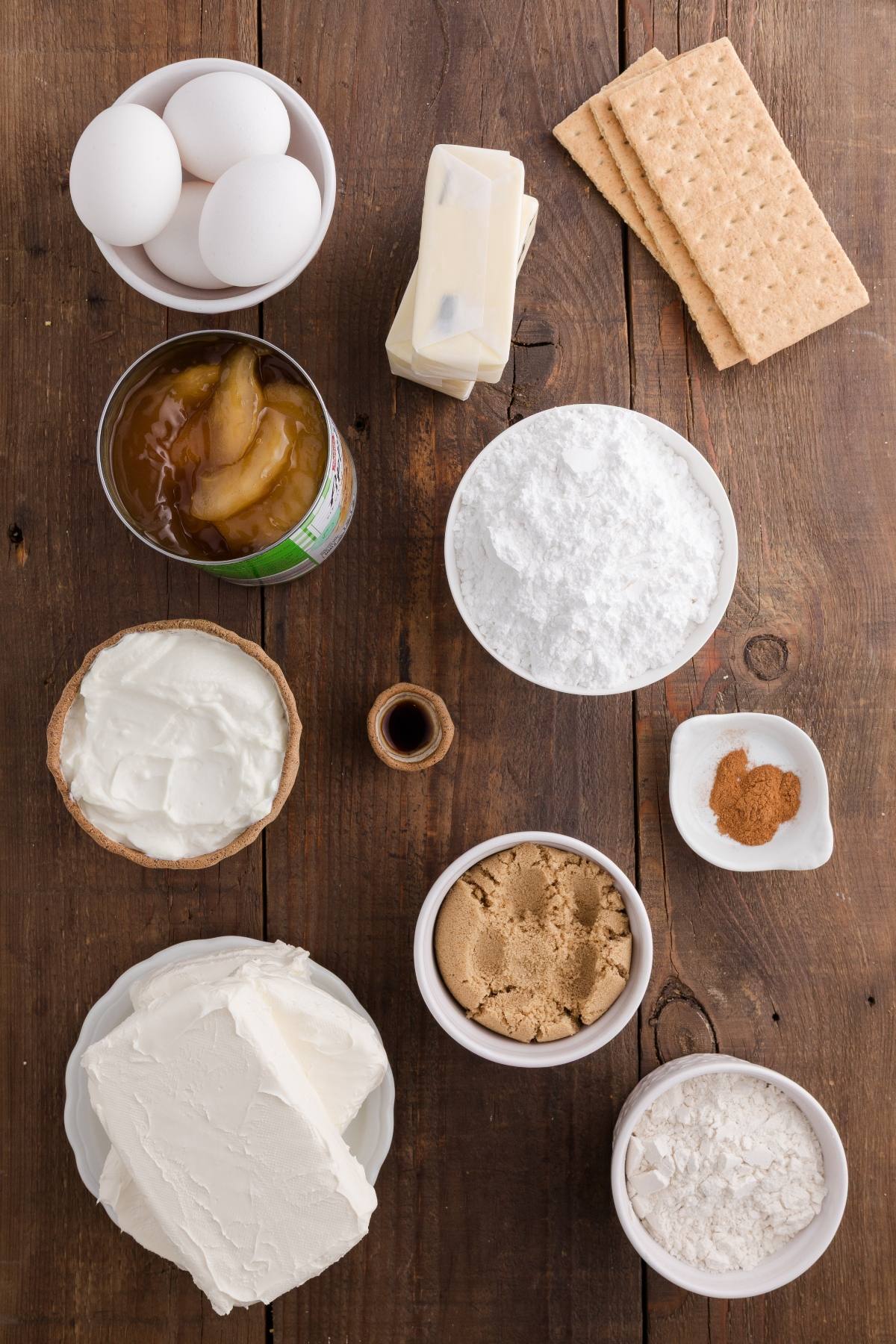 Baking ingredients on a wooden table: eggs, butter, graham crackers, cream cheese, sugar, flour, and canned apples.