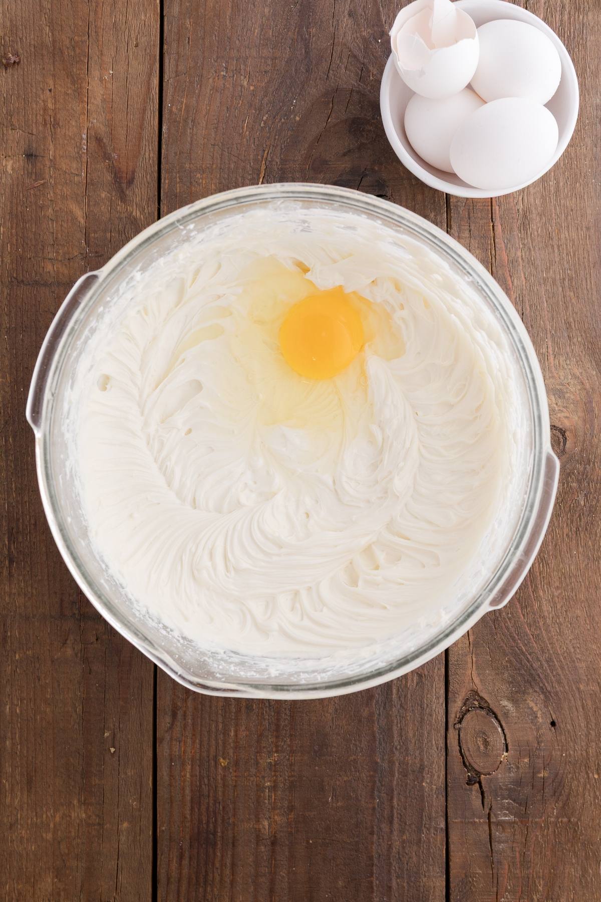A glass bowl of cream cheese mixture with an egg on top, next to a bowl of whole and cracked eggs.