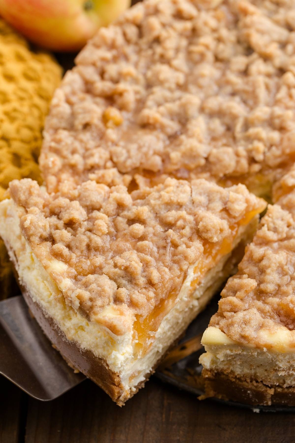 A slice of crumb-topped apple cheesecake being served from a whole cheesecake on a wooden table.