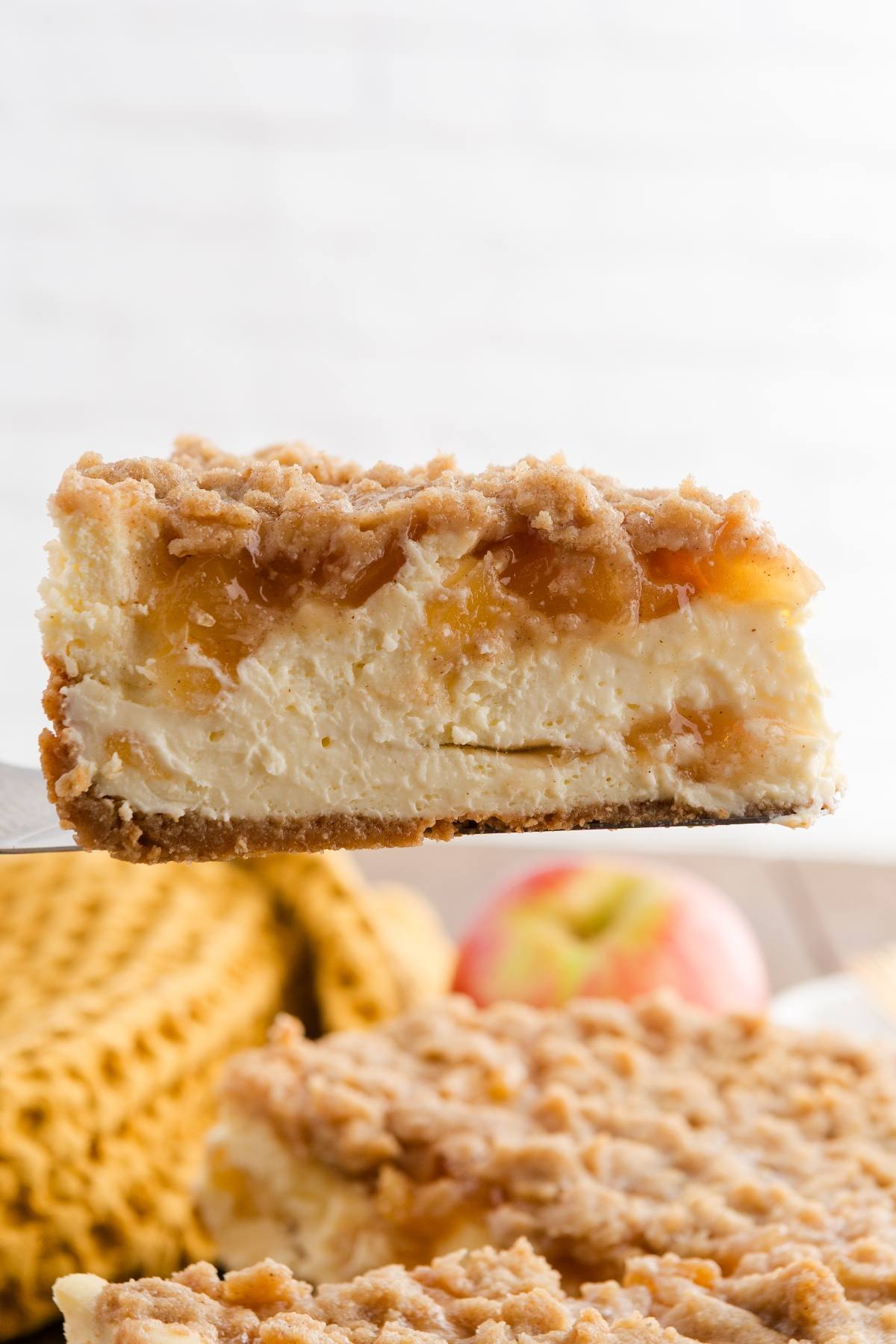A slice of crumb-topped cheesecake with apple filling held above a whole cheesecake, with an apple in the background.