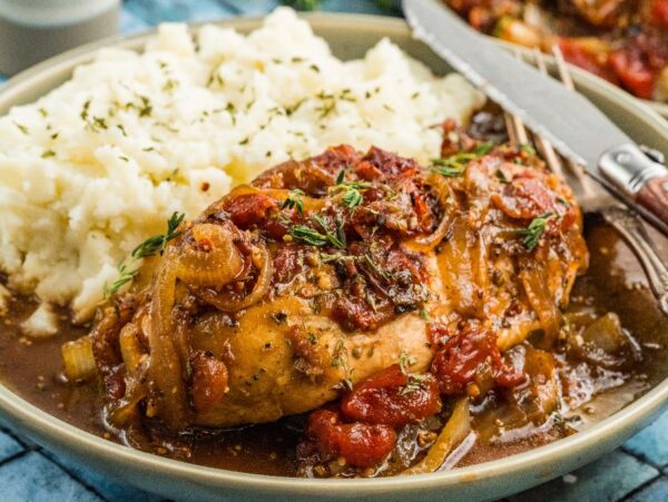 A plate of balsamic chicken smothered with gravy, tomatoes, and onions, served alongside mashed potatoes.