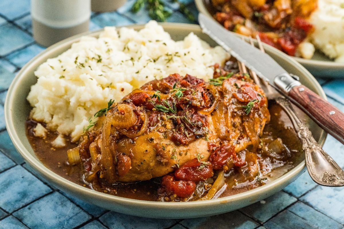 Plate of mashed potatoes and balsamic chicken braised with tomato sauce and herbs, served with a fork and knife.